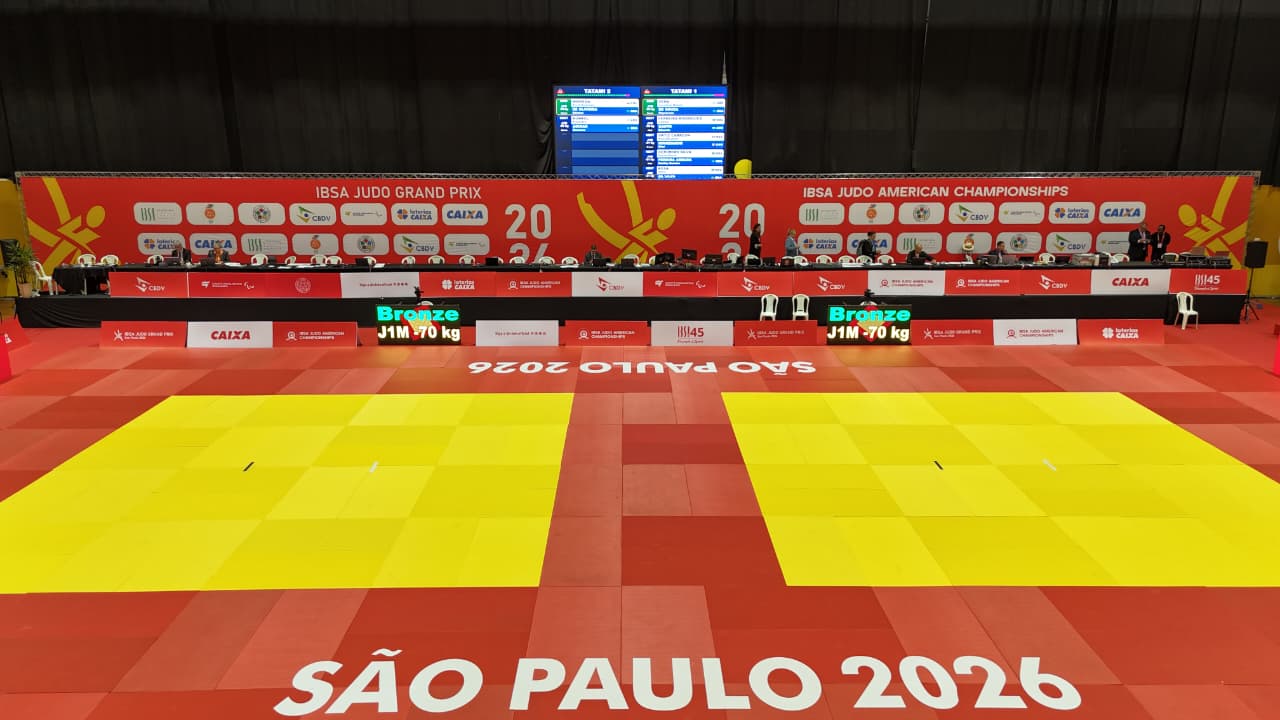 Vista panorâmica do ginásio que sediou o IBSA Judo Grand Prix e o IBSA Judo Americas and Oceania Championships São Paulo 2026. Em primeiro plano, aparecem duas áreas oficiais de competição de judô, com tatames amarelos cercados por áreas de segurança vermelhas. Ao fundo, está a mesa principal da organização, equipada com computadores, monitores e sistema de arbitragem, além de um grande painel vermelho com a identidade visual oficial dos eventos e os logotipos da IBSA, CBDV, Governo do Estado de