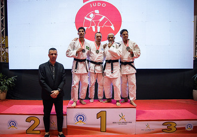 Em primeiro plano, à esquerda da imagem, está Felipe Menescal, Gerente Técnico de Eventos da CBDV, vestindo terno escuro e posando em frente ao pódio. Ao fundo, sobre o lugar mais alto do pódio, estão os quatro medalhistas da categoria. Da esquerda para a direita: o brasileiro Gabriel F. Rodrigues, medalha de prata; o argentino Eduardo Gauto, campeão da categoria com a medalha de ouro; o brasileiro Deyverson de Souza, medalhista de bronze; e o brasileiro Elielton de Oliveira, também medalhista d